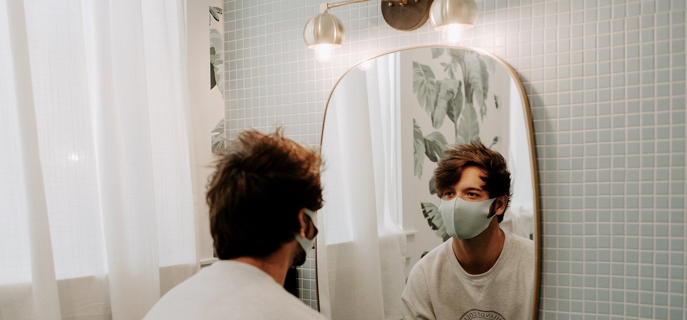 Mann steht in einem Badezimmer und betrachtet sich im Spiegel. Er trägt dabei eine Mundschutzmaske.