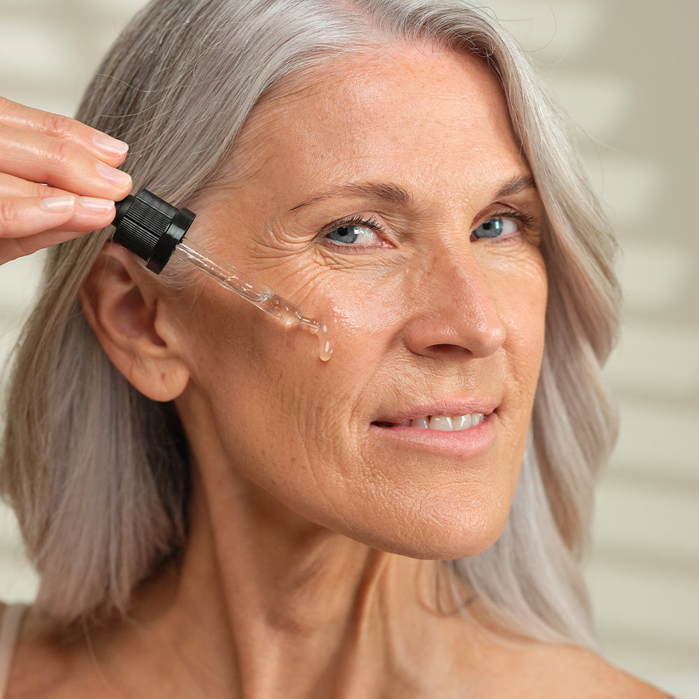 Eine Frau mit grauem Haar tröpfelt ein Serum aus einer Pipette auf ihr Gesicht und lächelt sanft.