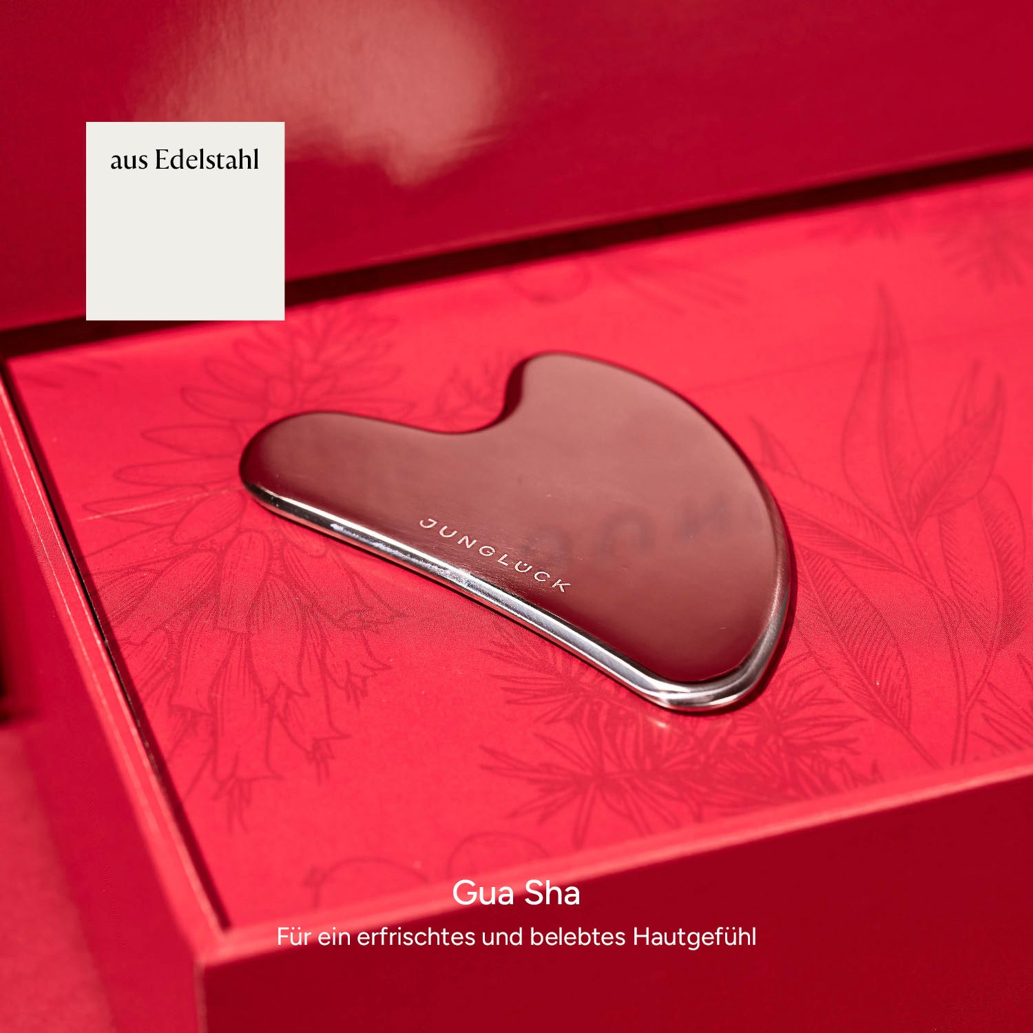 Ein Gua Sha aus Edelstahl liegt in einer roten Verpackung. Die Oberfläche reflektiert Licht und wirkt ansprechend.