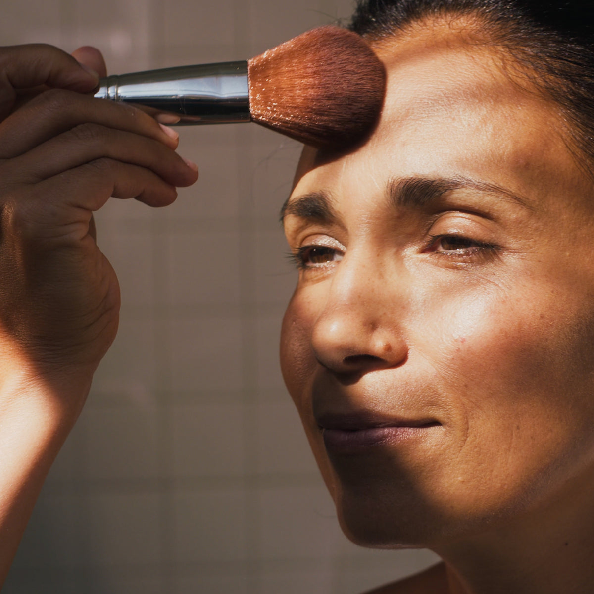 Eine Frau trägt mit einem Pinsel Transparent Puder auf ihre Stirn auf. Weiche Lichtakzente betonen ihre Gesichtszüge.